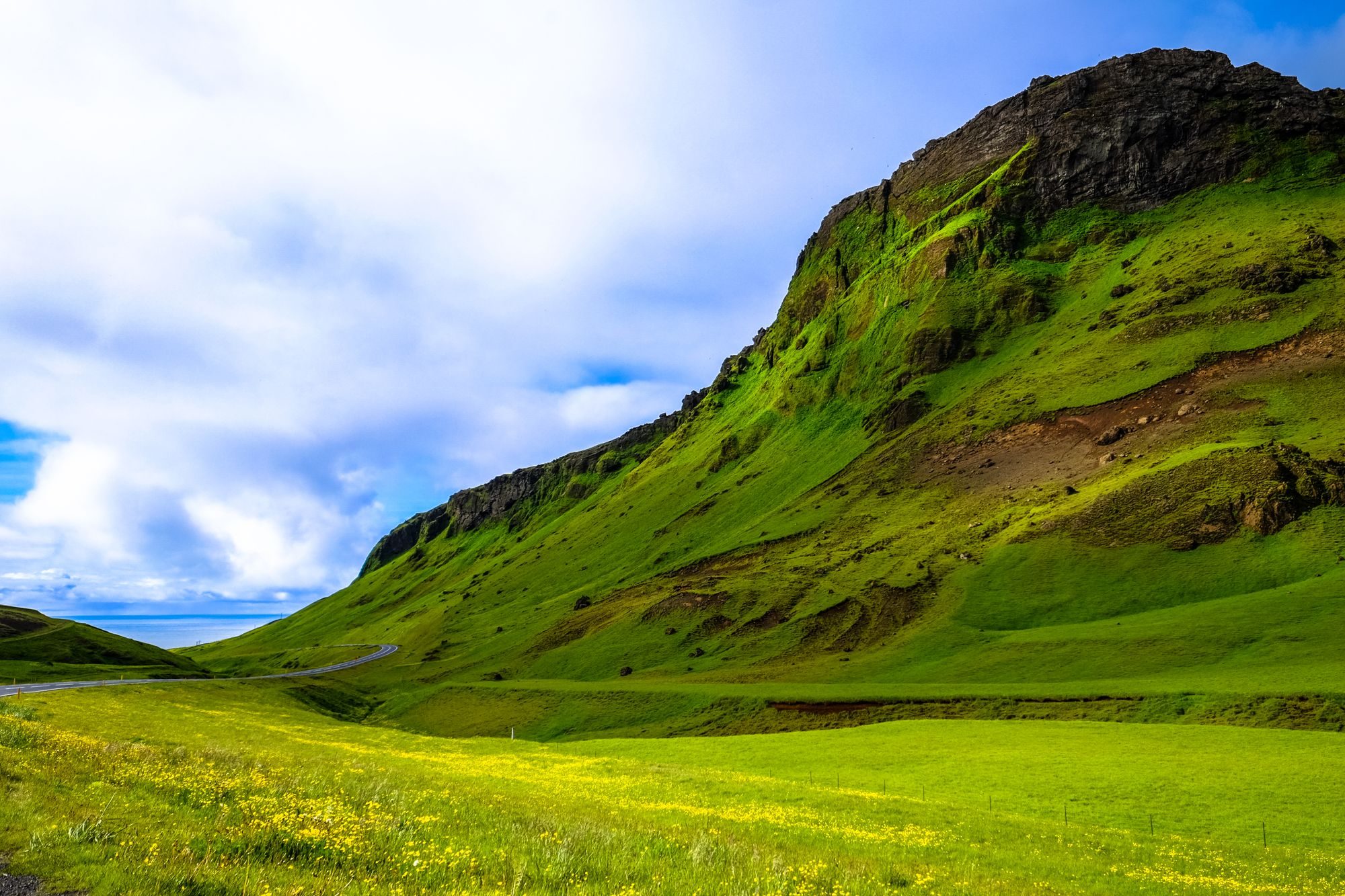 Green mountain slope above an open meadow beneath a blue sky and scattered clouds